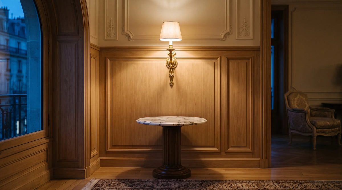 Applique de laiton allumée contre une boiserie claire, au-dessus d'un guéridon de marbre laissé vide. Image