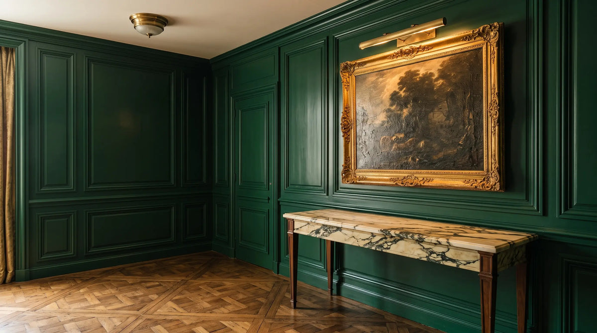 Tableau ancien à cadre doré éclairé par une applique cimaise de laiton, sur un mur de boiserie laquée vert profond, au-dessus d'une console de marbre veiné.
